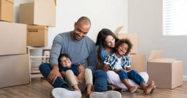Happy family with two children having fun at new home. Young multiethnic parents with two sons in their new house with cardboard boxes. Smiling little boys sitting on floor with mother and dad.