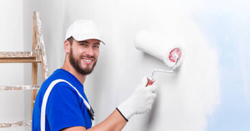 Portrait of handsome young smiling painter in white dungarees, blue t-shirt, cap and gloves painting a wall with paint roller, looking at camera