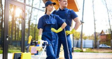Two friends from cleaning service busy with house cleaning session, stand closely outdoors, cleaning cottage, country house. Hygiene concept.